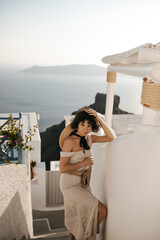Fototapeta na wymiar Brunette lady in beige midi dress touches hair. Curly charming woman in stylish outfit with straw bag poses in Greek town with sea view.