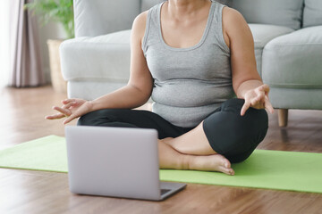 Fototapeta premium Mature Asian chubby fat woman sitting on the floor in living room practice online yoga lesson with the computer. female having meditate training class on the laptop..