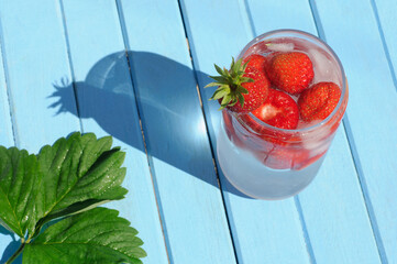 Summer cocktail made from fresh strawberries and ice on a blue wooden table with strawberry leaves. The shadow on the table from a glass with a drink