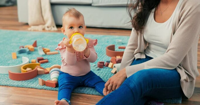 Sweet girl is sitting on carpet among toys with mother, holding bottle, cup in hands, drinking, gives it to woman's mouth to drink with her, she turns cup to see if water will spill out