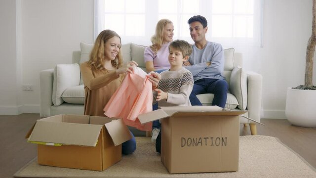 Cute little caucasian boy putting things in cardboard box for donation with help of teenage sister while happy mother and father watching them at home
