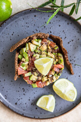 Raw tuna and avocado tartar served with rye bread crackers and lime on black ceramic plate. Gourmet fish tartar.