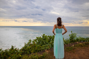 Fototapeta na wymiar Travel lifestyle. Young woman enjoying ocean view and wearing long dress. View from back. Horizon seaview. Cliff near Balangan beach. Copy space. Bali, Indonesia