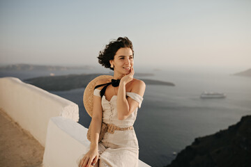 Brunette short-haired woman in elegant beige dress and boater enjoys beautiful sea view. Curly lady in good mood sits on white fence.