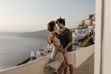 Fototapeta na wymiar Portrait of couple on background of Greek city. Brunette lady in beige dress and man in stylish outfit looks at each other with love on sea background.