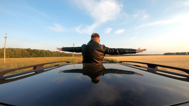 Young guy standing out of car sunroof with hands to sides and flying like plane while riding through country road. Man enjoying road trip and wind during journey. Travel or freedom concept. Back view