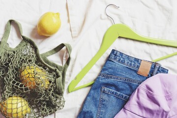 Flat lay composition photo items on a bed. Trendy stylish blue jeans, lilac panama hat, green hanger and cotton bag shopper with lemons. Top view fashion object photography