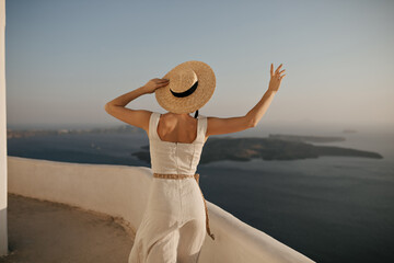 Charming tanned woman in beige dress and straw hat walks on sea background. Lady in elegant outfit...