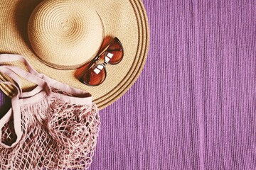 Trendy summer beach accessories for women. Straw hat, elegant brown sunglasses and fishnet cotton bag on a purple mat. Flat lay fashion photography