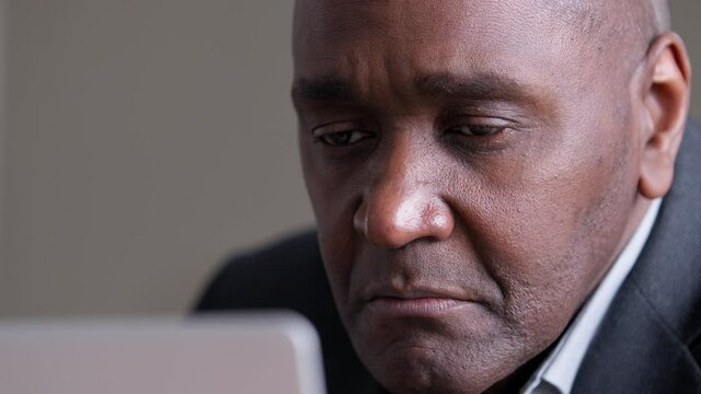 Close-up focused male wrinkled face looking at laptop screen, african mature business man uses computer in office reading news email online emotionally raising eyebrows, 40s afro boss working on net