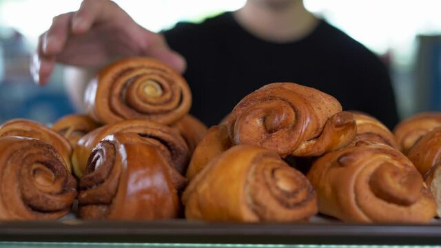 Mouth-watering, large, brown, homemade cinnamon rolls are stacked in a grocery store window at a bakery. The man's hand takes two.