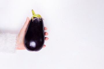 The hand of a young girl in a white fluffy sweater holds a ripe whole eggplant on a white background copy space