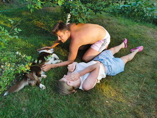  A young man and a woman on the grass, stroking and kissing a brown Welsh corgi cardigan dog.