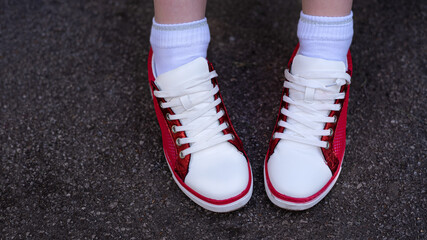 Photo of female legs in red and white sneakers on gray wet asphalt. Red and white sneakers with white laces close up