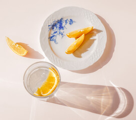 Sliced orange on white vintage plate with glass of water and sharp shadows stands on light pink...