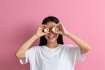 Asian beauty model girl eating Sushi roll, healthy japanese food. Beautiful woman holding Philadelfia roll near her eyes in hands like binoculars and looking for tasty sushi