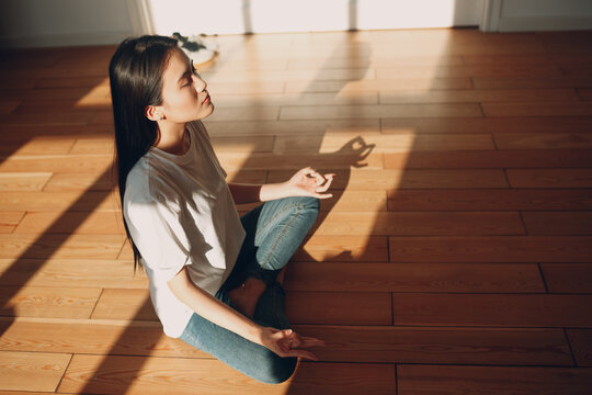 Asian woman doing yoga and zen like meditation lotus pose in casual wear at indoor living room apartment with natural sun light with shadows