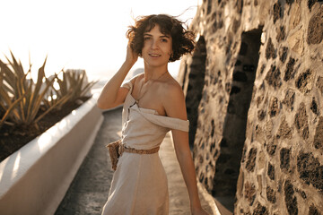 Curly brunette lady in stylish beige dress with straw bag moves along path in old city and smiles...