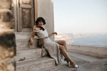 Short-haired brunette woman in midi dress looks into camera and sits on stairs near old house with sea view. Charming lady in summer outfit poses near wooden door.