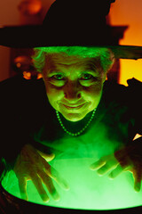 Smiling old woman in classic witch costume preparing green magic potions and looking at camera. senior person celebrating halloween