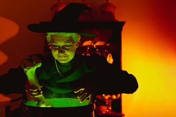 Older woman disguised as a classic witch preparing magic potions in a copper pot that emits green...