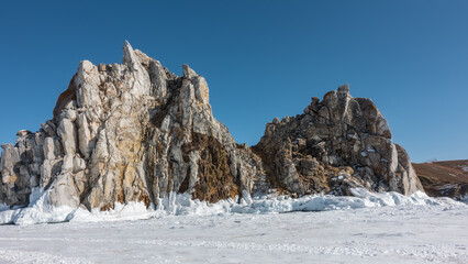 A two-headed rock, devoid of vegetation, rises above a frozen lake. Cracks on the rocks, a thick layer of icicles on the base of the cliff. Snow on ice. Clear blue sky. Baikal