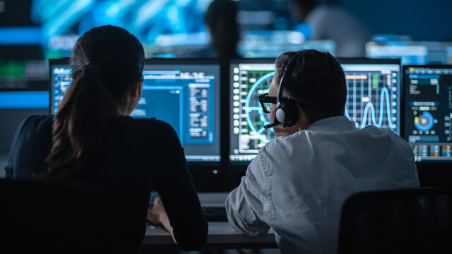 Two Specialist Work on a Computer with Live Ananlysis Feed from a Global Map on a Big Digital Screen. Employees Sit in Front of Displays with Financial Stock Market Trading Info and Big Data.
