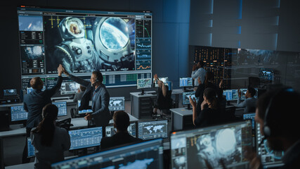 Group of People in Mission Control Center Establish Successful Video Connection on a Big Screen with an Astronaut on Board of a Space Station. Flight Control Scientists Sit in Front Computer Displays. - Powered by Adobe