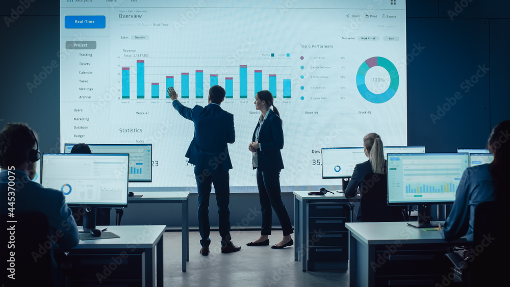 Aufkleber Two Traders Having a Meeting in a Modern Monitoring Office with Analytics Feed on a Big Digital Screen. Monitoring Room with Brokers and Finance Specialists Sit in Front of Computers. Colleagues Talk.