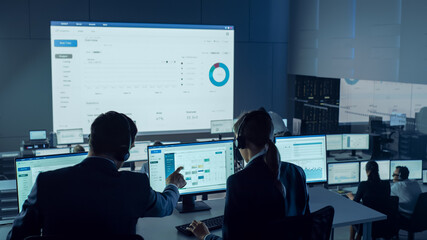 Professional Traders Working in a Modern Monitoring Office with Live Analytics Feed on a Big Digital Screen. Monitoring Room with Brokers and Finance Specialists Sit in Front of Computers. 