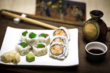 A plate with maki sushi rolls made of rice, raw fish, green caviar, sesame seeds, nori and fried tempura prawn with wasabi, pickled ginger, soy sauce and wooden chopsticks, with asian decoration