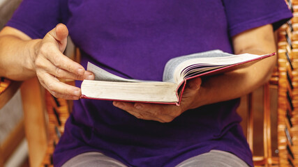 An elderly woman is reading a book, holding it in her hands and sitting in a chair. Reading the Bible