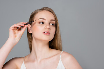 young woman applying brown eyebrow pencil and looking away isolated on grey