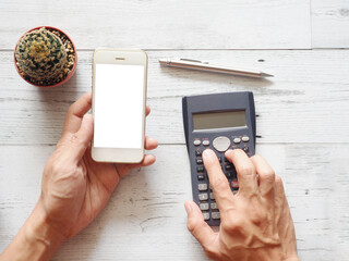 Fototapeta na wymiar Hand holding cellphone white screen and using calculator with cactus and silver pen on white wood table nature shadow and sunlight top view and space business concept