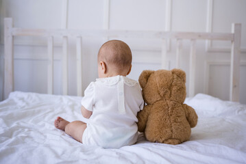 baby girl is sitting with her back on the bed with a teddy bear toy © Яна Айбазова