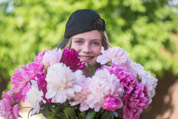 Beautiful and happy young girl in a cap and sunglasses in summer with a beautiful bouquet of peonies in nature