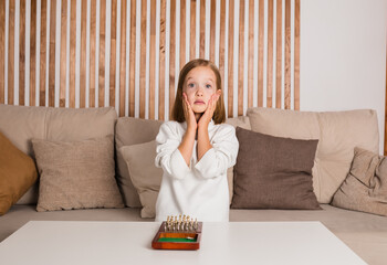 shocked little chess player in a white pullover is sitting on the sofa and playing chess