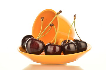 Several juicy berries of dark red cherries on a yellow saucer with a ceramic cup, close-up, isolated on white.