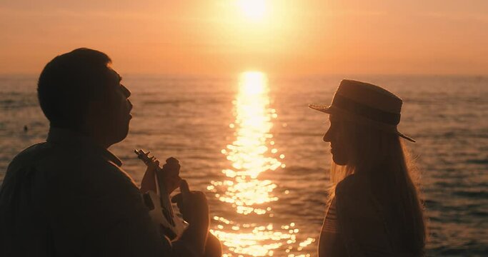 Fun attractive couple of traveler sitting on sea beach with ukulele under sunset sky in evening time. Man playing, woman listening and sing music, relaxing enjoying holidays, travel day. 4K video