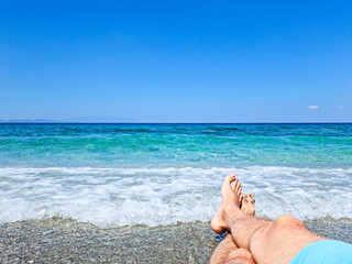 Sea wave washes the feet of a man sitting on the sand of the beach. Blue sea and sky. Relax concept