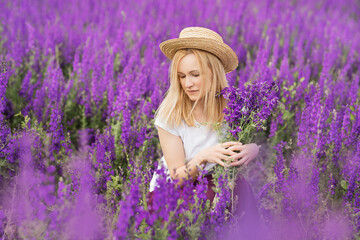 A girl in a straw hat with a bouquet of delphiniums is resting in a field. Spring day in a blooming field of delphiniums