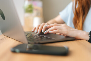 Close-up hands of unrecognizable young woman typing text on laptop keyboard sitting at table by window in cafe. Selective focus of female working on laptop on background of blurred window in daylight.