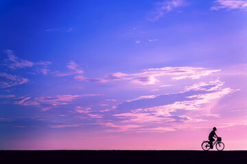 Naklejka na ściany i meble The silhouette of a cyclist at dawn. A man rides a bicycle with a basket on the background of a purple sky. Cyclist in Bali