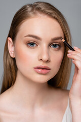 young woman with lip gloss applying brown eyebrow pencil isolated on grey