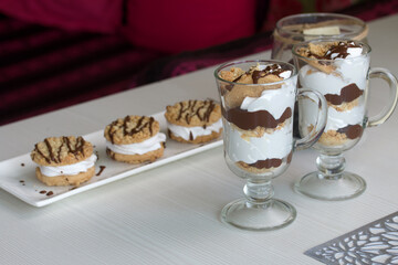 Dessert of biscuits, cream and chocolate. Next to it is a biscuit and cream sandwich. Decorated with chocolate. Lies on a plate.