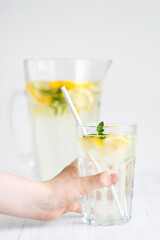 Child hand holding glass of lemonade against pitcher of lemonade on the white table.