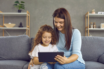 Cheerful young mother and her cute little daughter use a tablet to watch a video or cartoon. Mom, sister or babysitter sitting with a little girl on the couch at home. Family weekend concept.