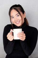Young oriental girl looks at the camera hugging with her hands a white cup.