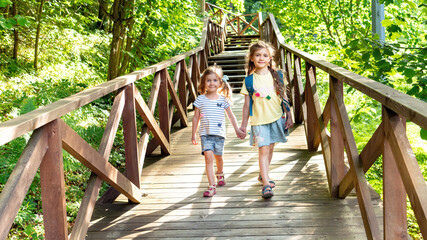 Kids with positive emotions travel on a hiking route to the reserve. Two smiling girls head to the family camping along a wooden road. Children's tourism concept. Happy facial expressions in kids.