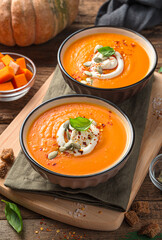 Two bowls of pumpkin soup with basil and cream on a wooden background.
